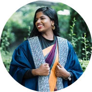 Pauline Suzanna smiling in a blue graduation gown and colorful traditional saree against a green foliage background.